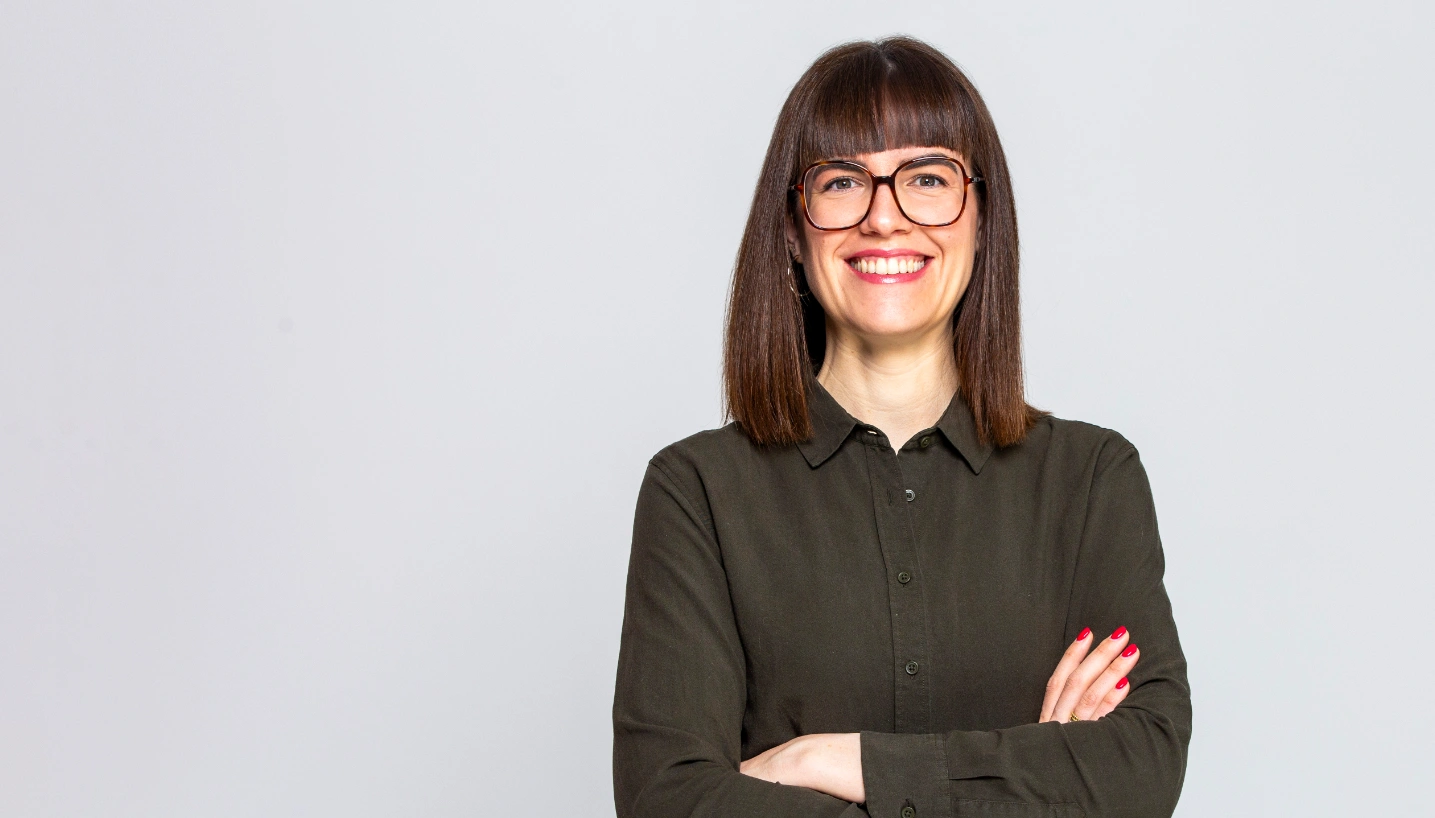 Frau mit dunklen Haaren und Brille, die in einem grauen Hemd lächelt und die Arme verschränkt.